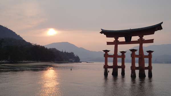 the miyajima torii gate with the sun starting to set in the background and reflected on the water.  the shore is visible on the left and there are mountains in the background